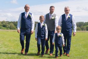The groom and groomsmen walking towards the camera, in an open field, with the sun shining.