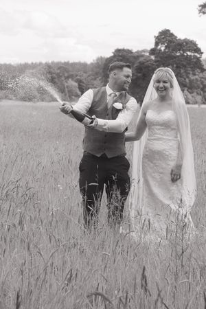 A black and white image of newlywed bride and groom, popping a bottle of champagne in the middle of a field. Bubbles flying everywhere