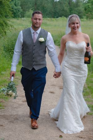 Bride and groom walking hand in hand. Groom is holding the wedding bouquet and the bride is holding a bottle of champagne.