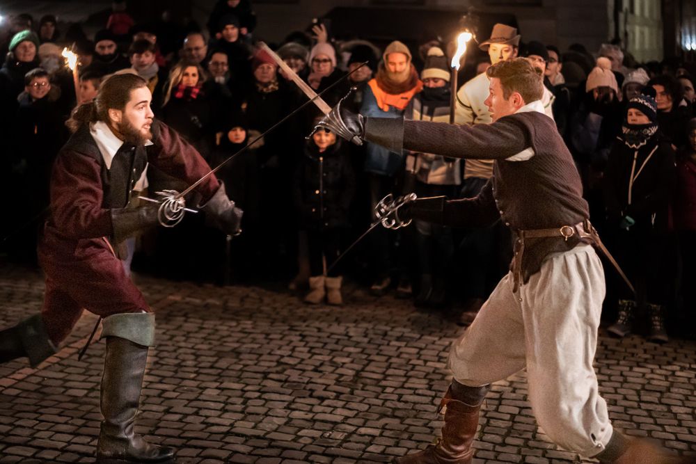 Two performers in historical costume duel with swords on cobblestone street as crowd watches.