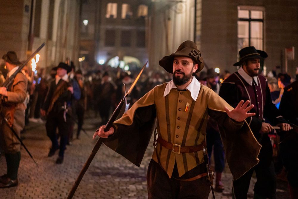Medieval festival participant in period costume carries a torch during nighttime celebration.