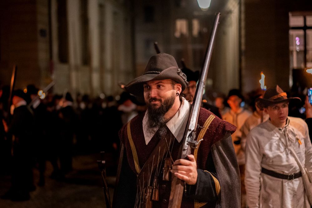 Historical reenactor in period costume holds a musket during a nighttime colonial demonstration.