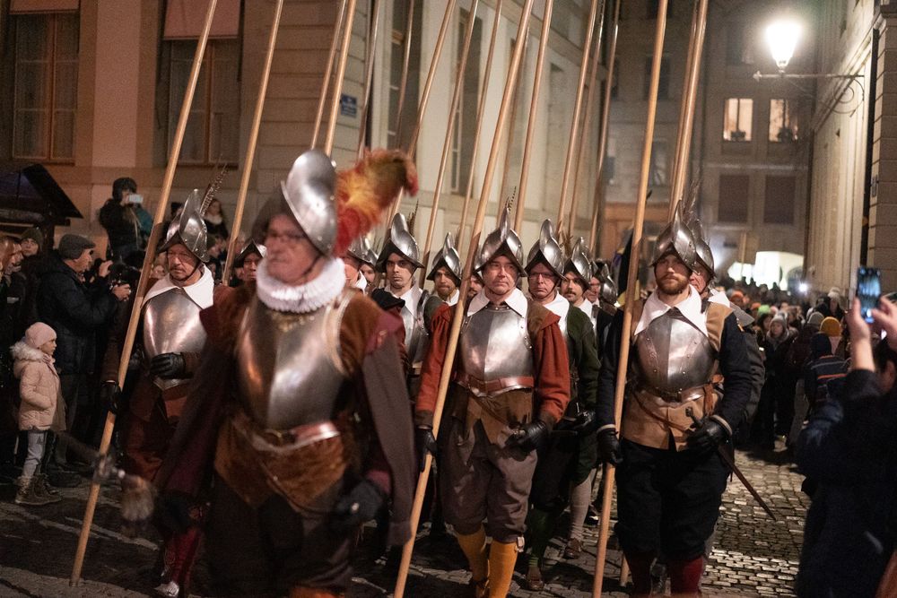 Medieval reenactors in armor carrying long spears march in formation at night.