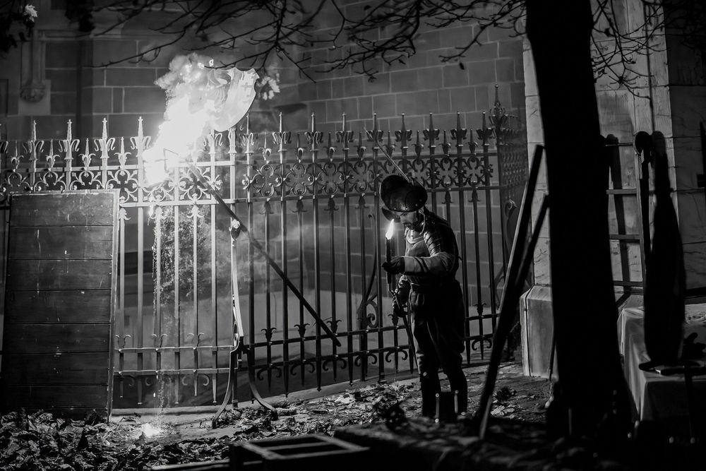 Person holding torch near iron gates at night in black and white.