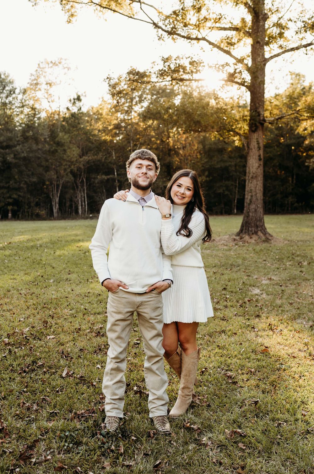 A couple in coordinating cream outfits poses together in a sunlit grassy field surrounded by autumn trees.