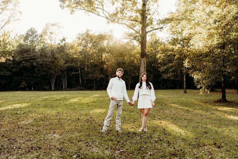 Couple holding hands in a sunlit green field surrounded by tall trees during a golden hour outdoor engagement session.