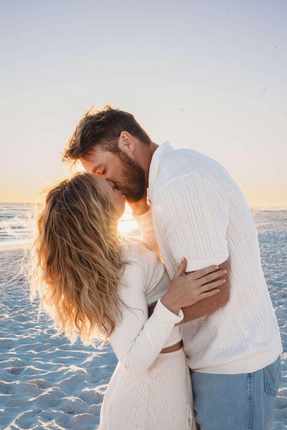 Romantic couple in white sweaters embrace on a beach at sunset, silhouetted against the golden light.
