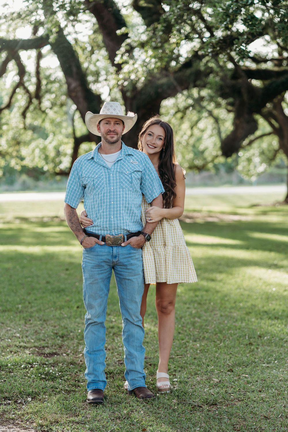A smiling couple poses outdoors in a park, man in cowboy hat and jeans, woman in yellow gingham dress.