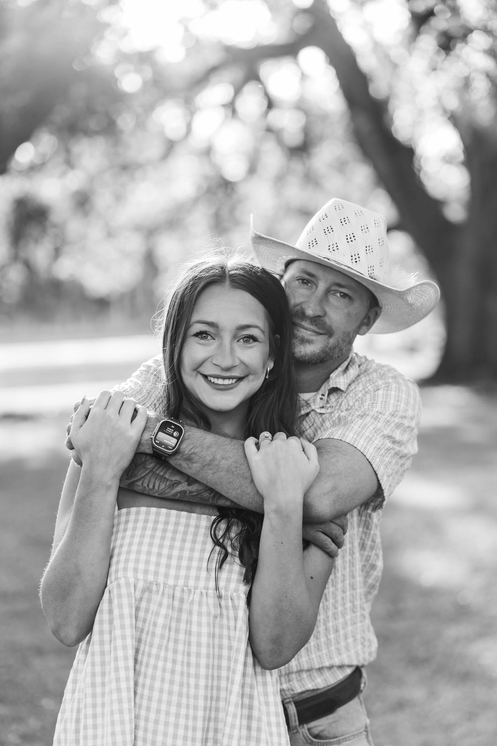 Black and white photo of a smiling couple embracing outdoors, man in cowboy hat hugging woman in gingham dress.