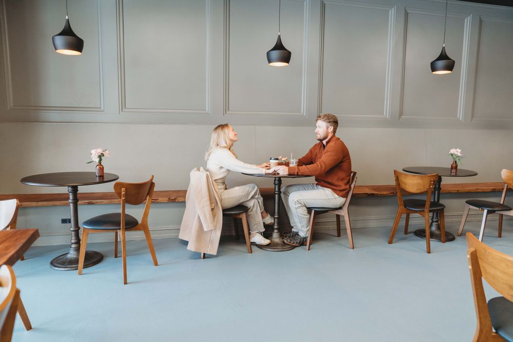 Two people sit at a small round table in a minimalist cafe with blue flooring and pendant lights overhead.