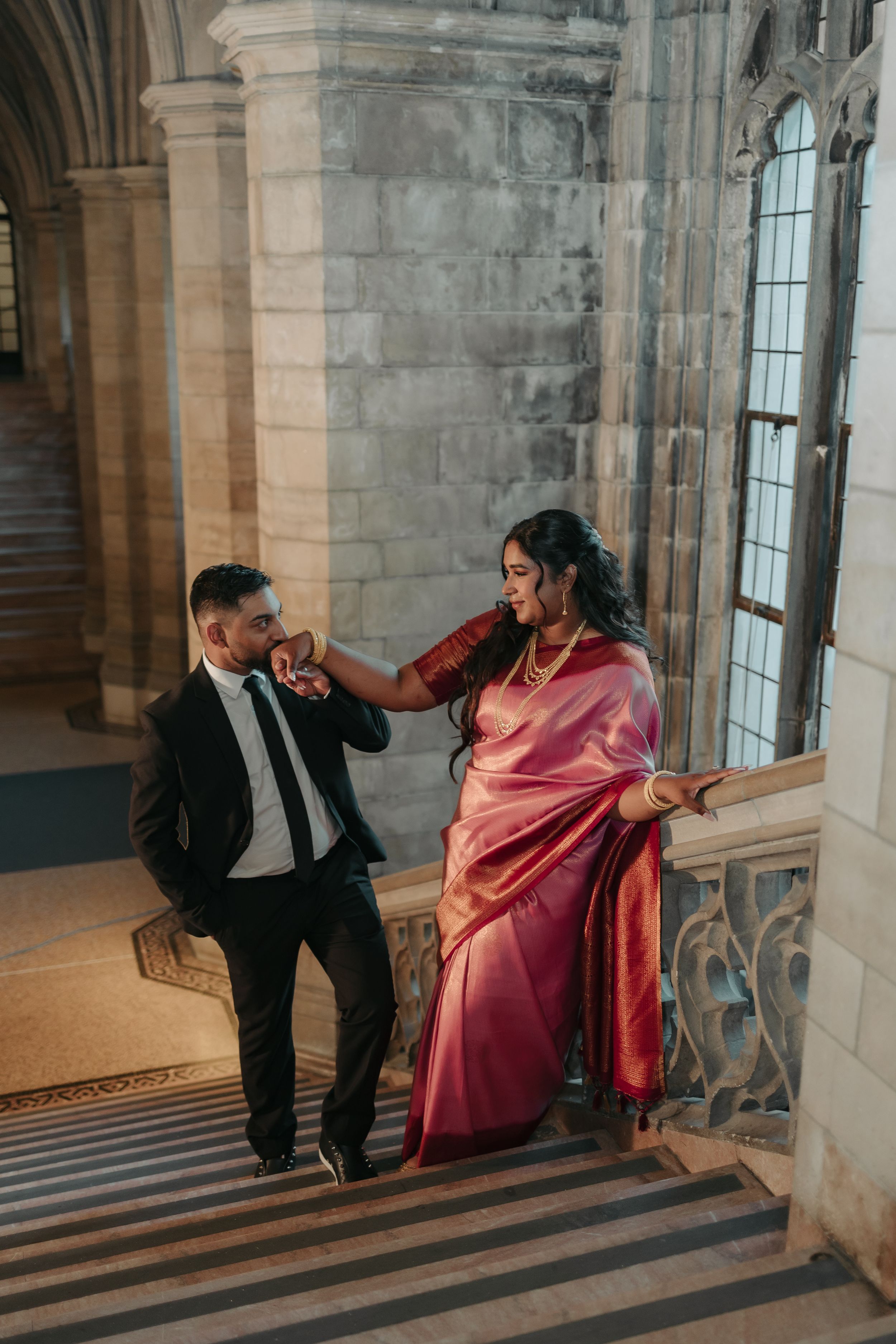 A couple in formal wear poses playfully on stone steps in a historic building.