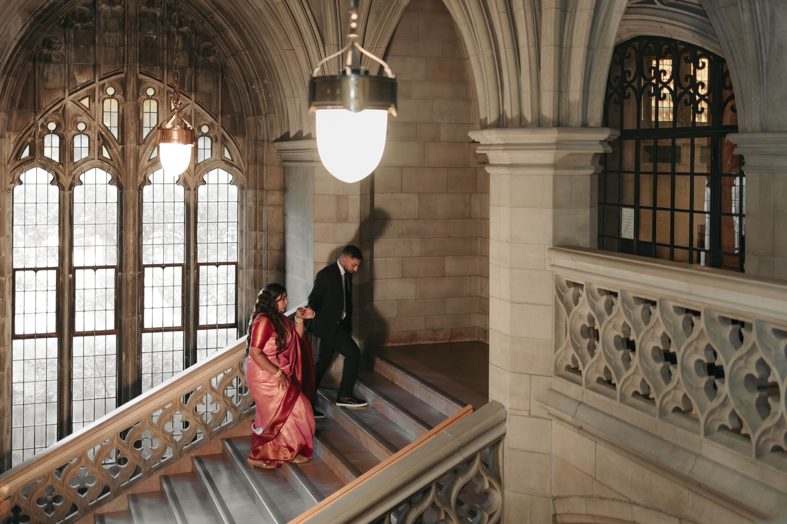 A couple ascends an ornate gothic stone staircase under pendant lighting.