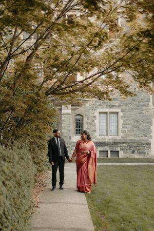 Couple walking hand in hand on path beneath autumn trees by stone building.