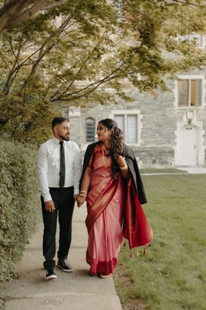 A couple strolls together on a garden path, with the person wearing a pink sari and black jacket beside stone architecture.