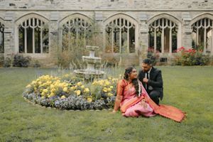 Two people sit together on grass near Gothic windows and a floral garden display with fountain.
