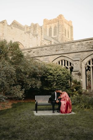 Two people share a quiet moment on a bench near Gothic church architecture and garden greenery.