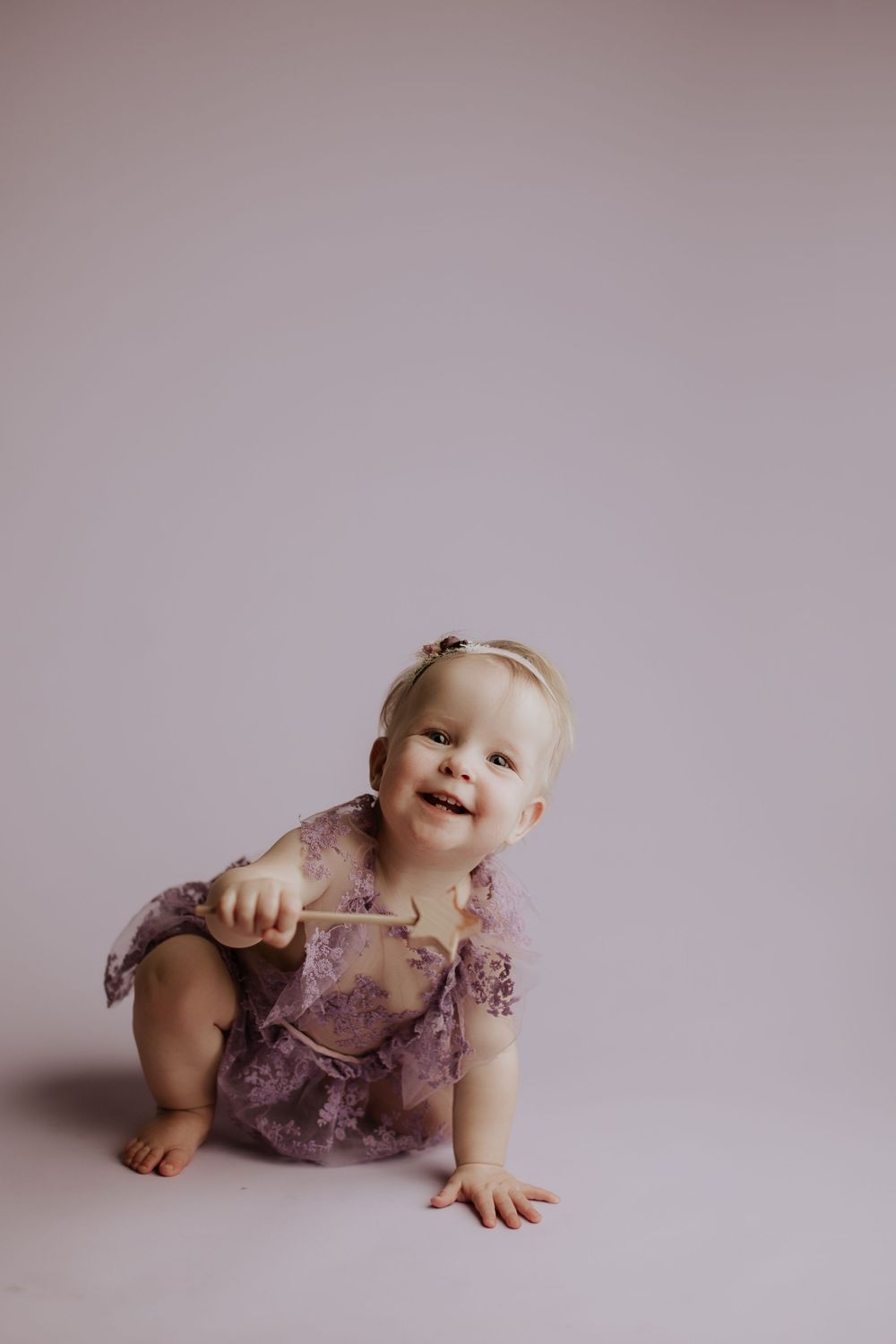 Happy baby girl in a purple floral lace romper crawling and smiling against a soft lavender background.