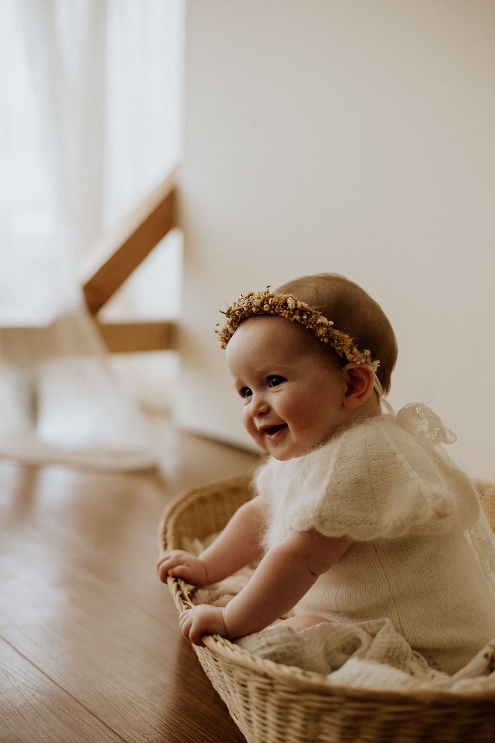 An adorable smiling baby wearing a floral crown and white dress sitting in a wicker basket.