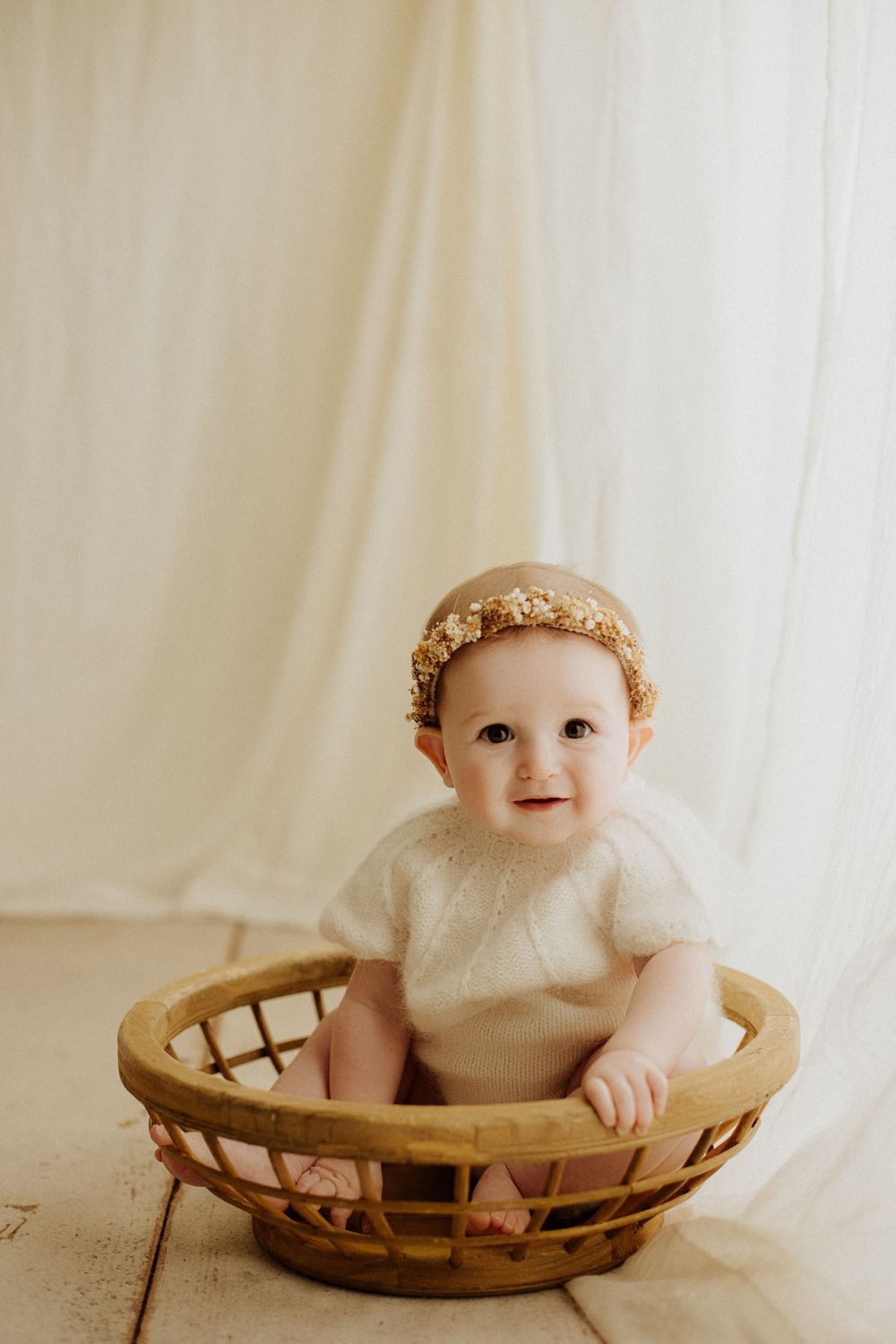 Adorable baby girl in a white knit romper and gold crown sitting inside a rustic woven wooden basket.