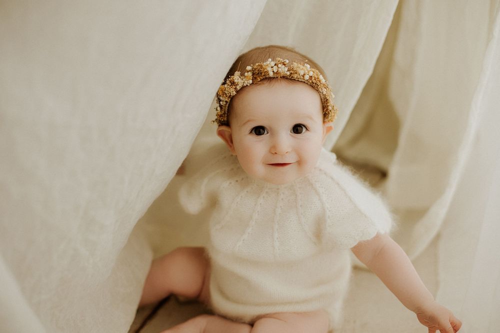 Smiling baby girl wearing a gold floral crown headband and white knit romper against a cream backdrop.