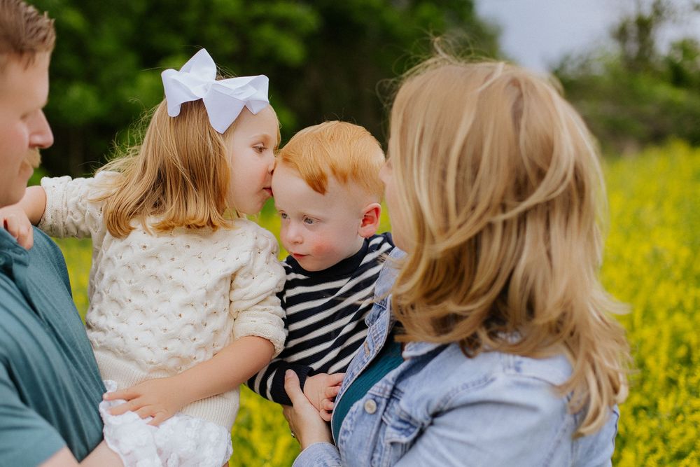 Young blonde girl with a white bow kissing her red-haired toddler brother during a family photo session.