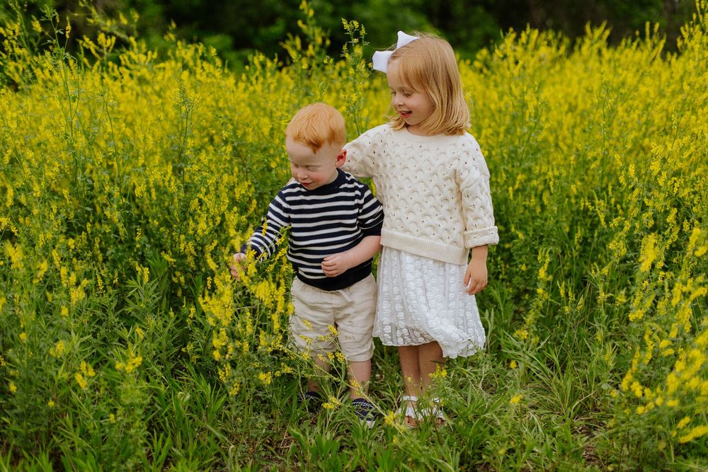 Sweet red-haired toddler boy and blonde girl in white dress standing together in a yellow wildflower field.