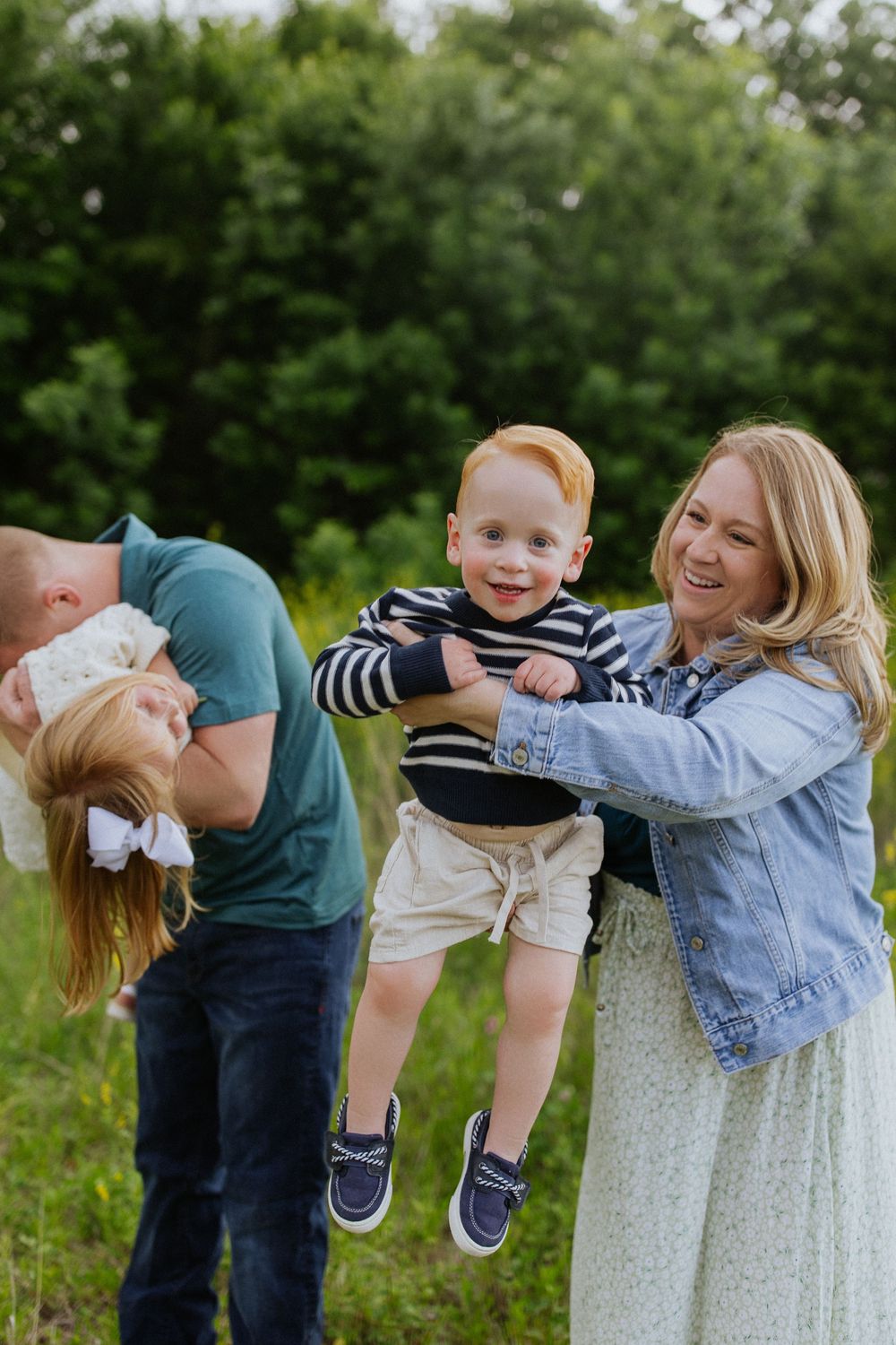 Joyful family playing outdoors as mom lifts smiling red-haired toddler while dad playfully dips daughter.