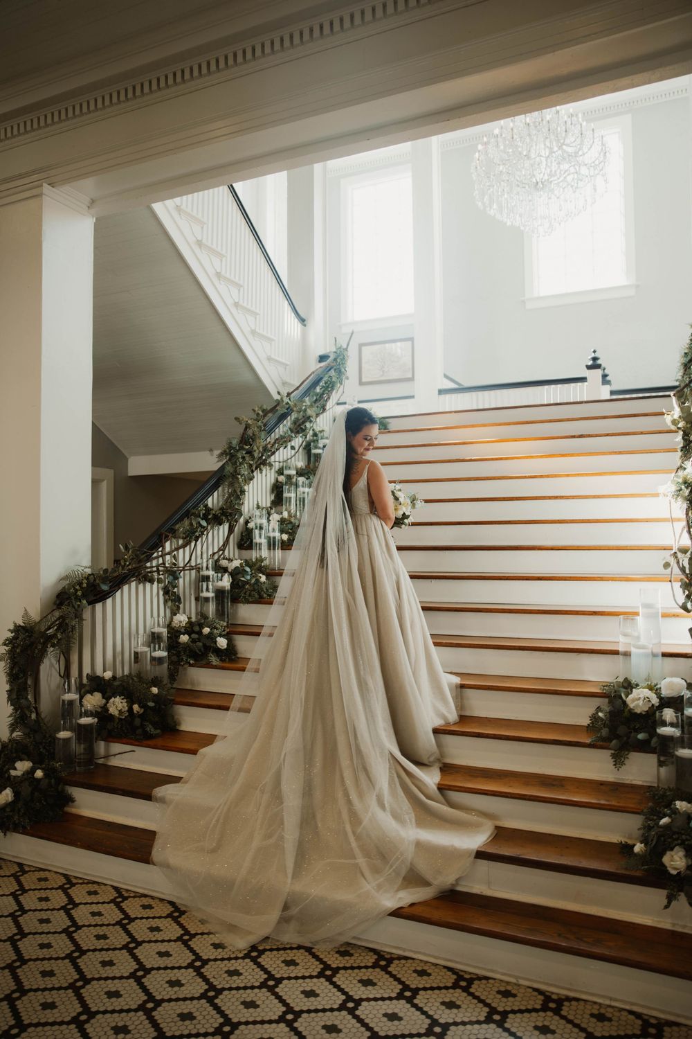 Elegant white wedding dress flowing down wooden stairs decorated with greenery and floral arrangements.