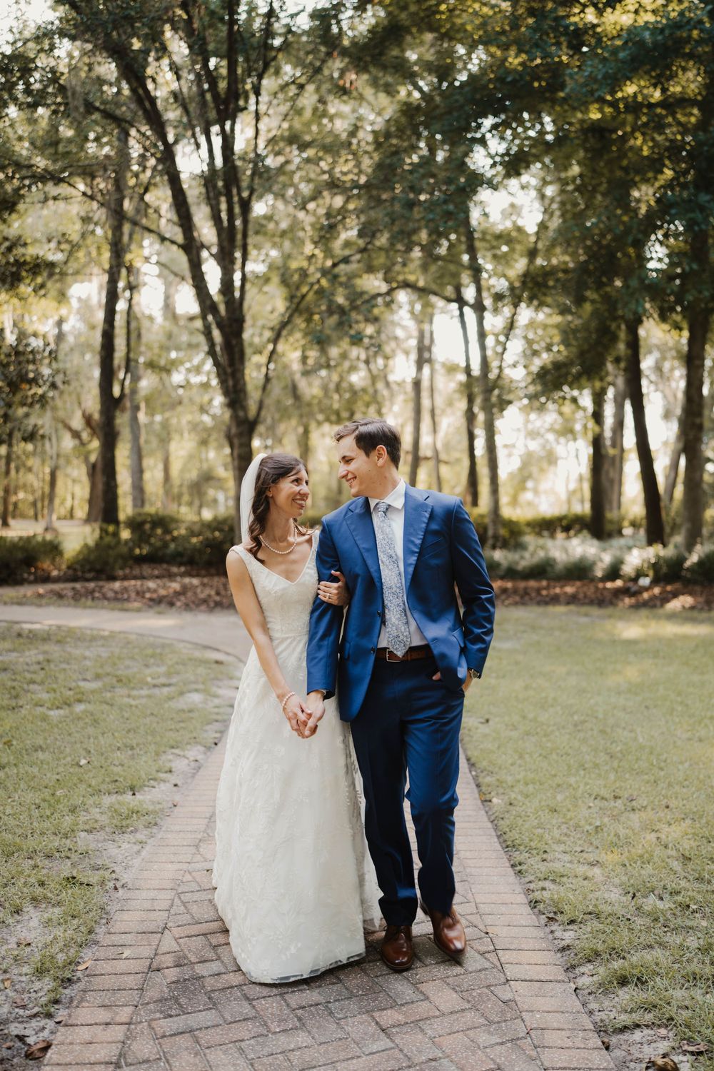 A wedding couple walks together along a brick path surrounded by trees.