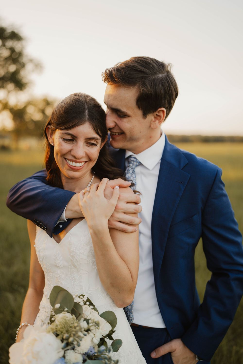 Newlyweds laughing together while holding white floral bouquet at outdoor wedding.
