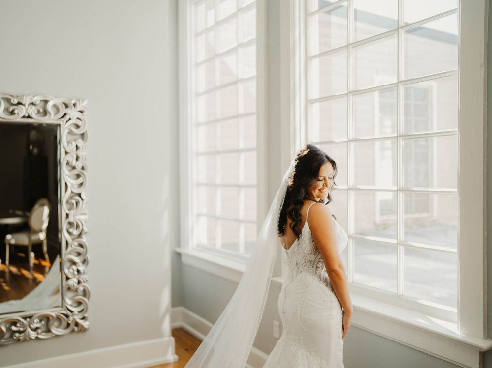 Bride in white dress poses by bright interior windows with ornate mirrors.