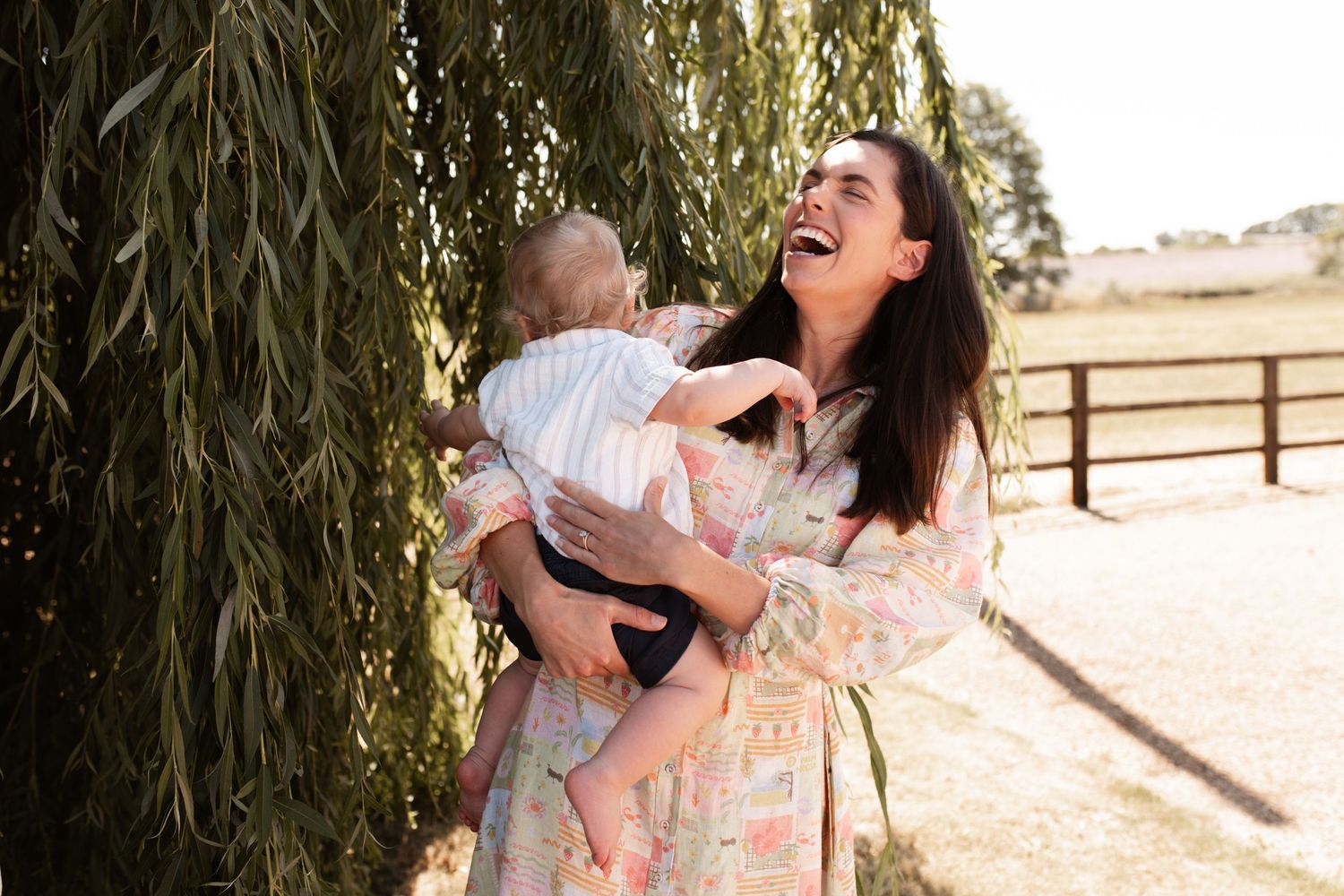 A joyful moment shared between parent and baby under a weeping willow tree on a sunny day.