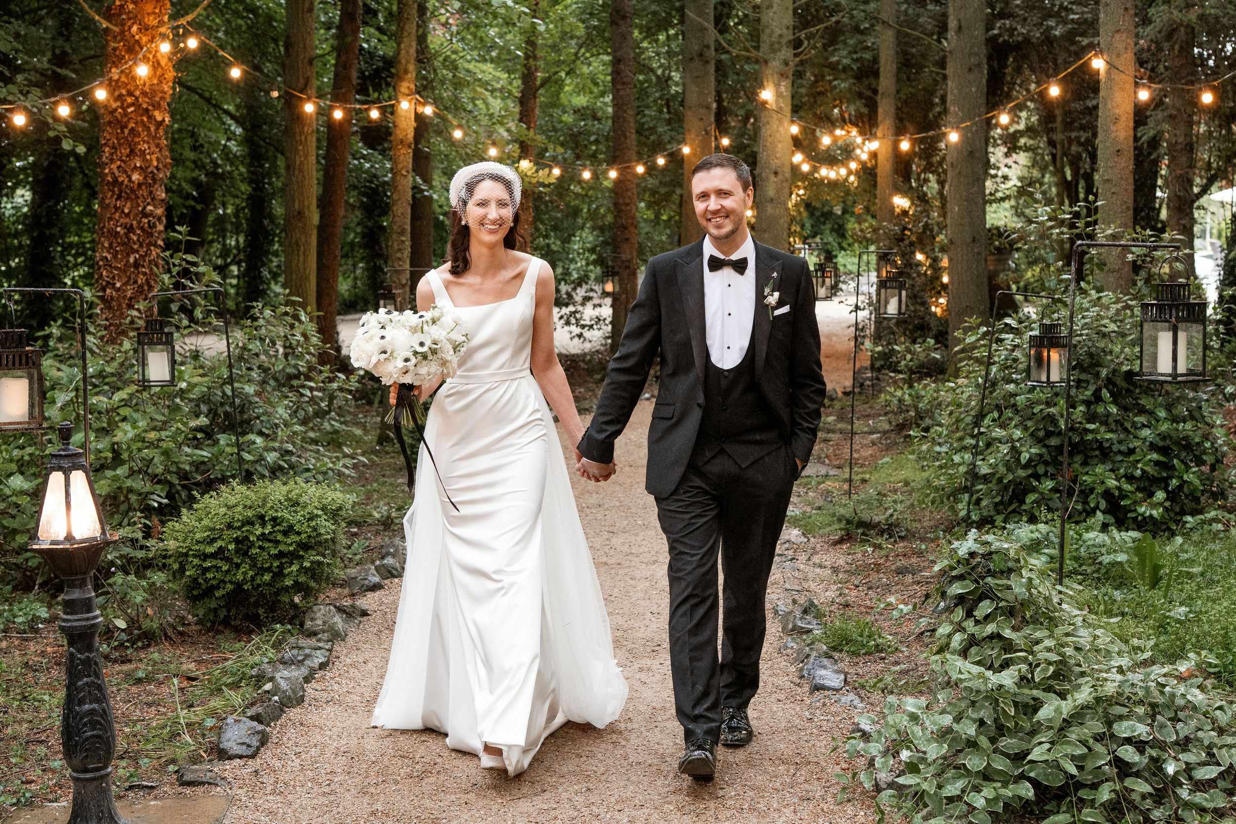 Wedding party walks along string lights decorated wooded path at outdoor reception.