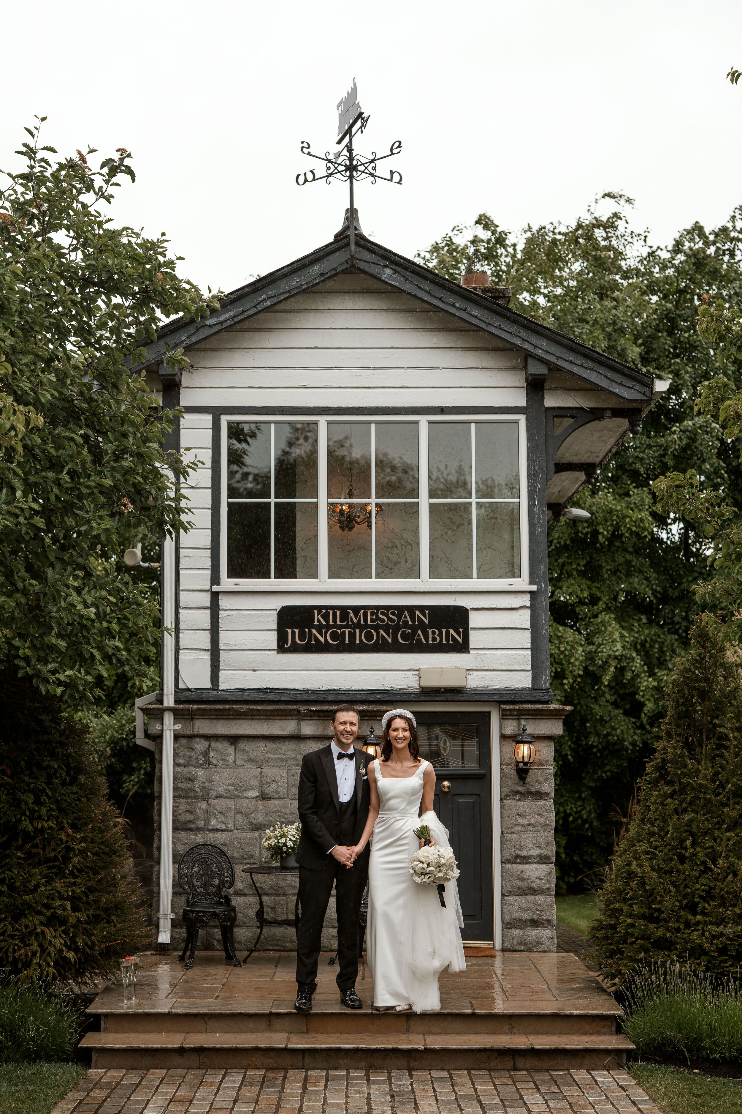 White rustic cabin venue with weathervane and stone foundation serves as wedding backdrop.