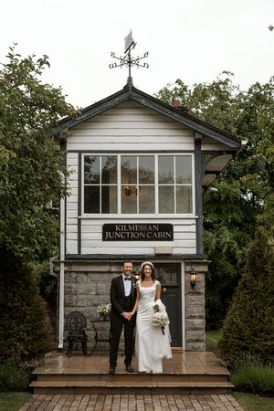 White rustic cabin venue with weathervane and stone foundation serves as wedding backdrop.