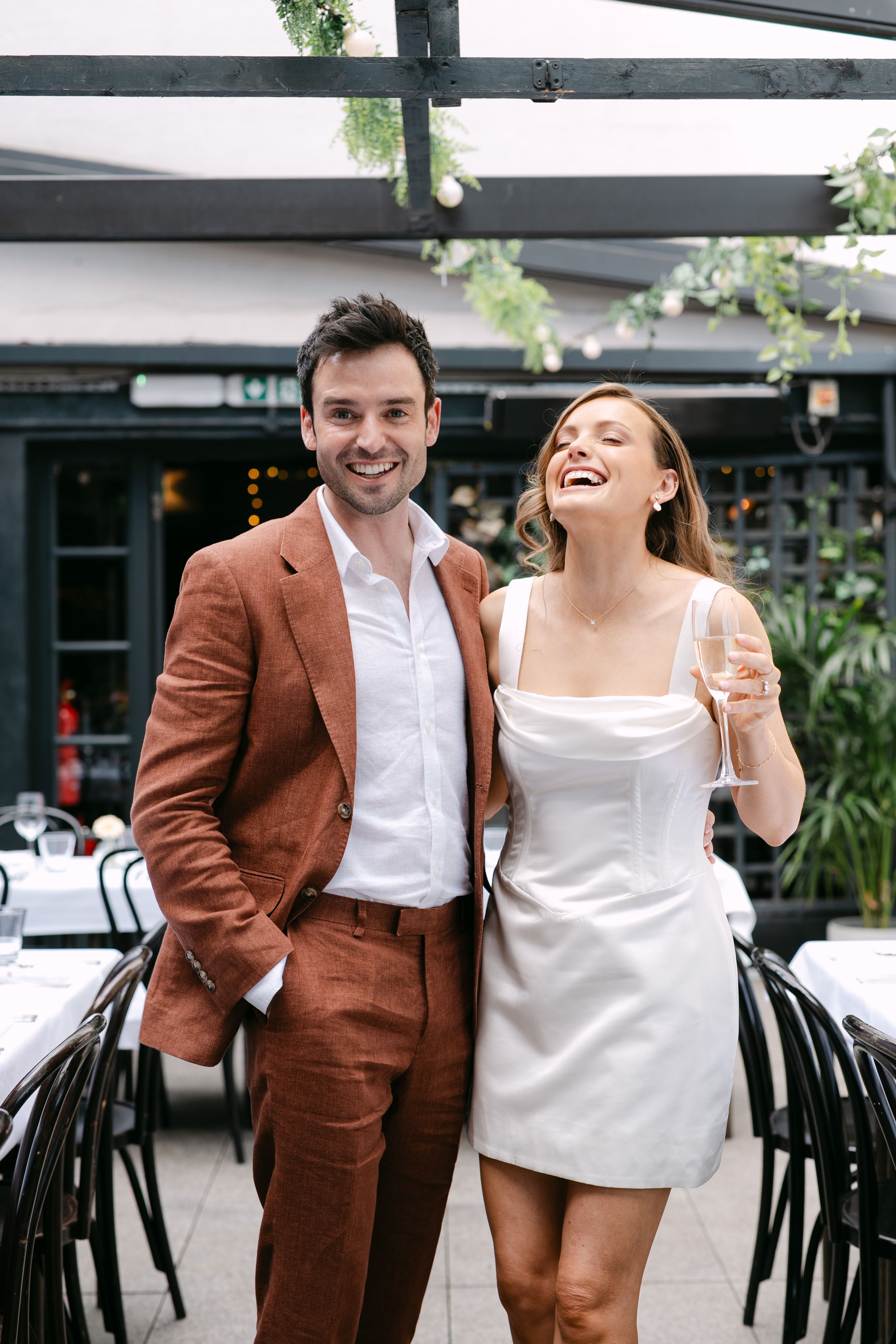 Smiling couple in elegant attire at a restaurant, man in brown suit and woman in white dress holding champagne.
