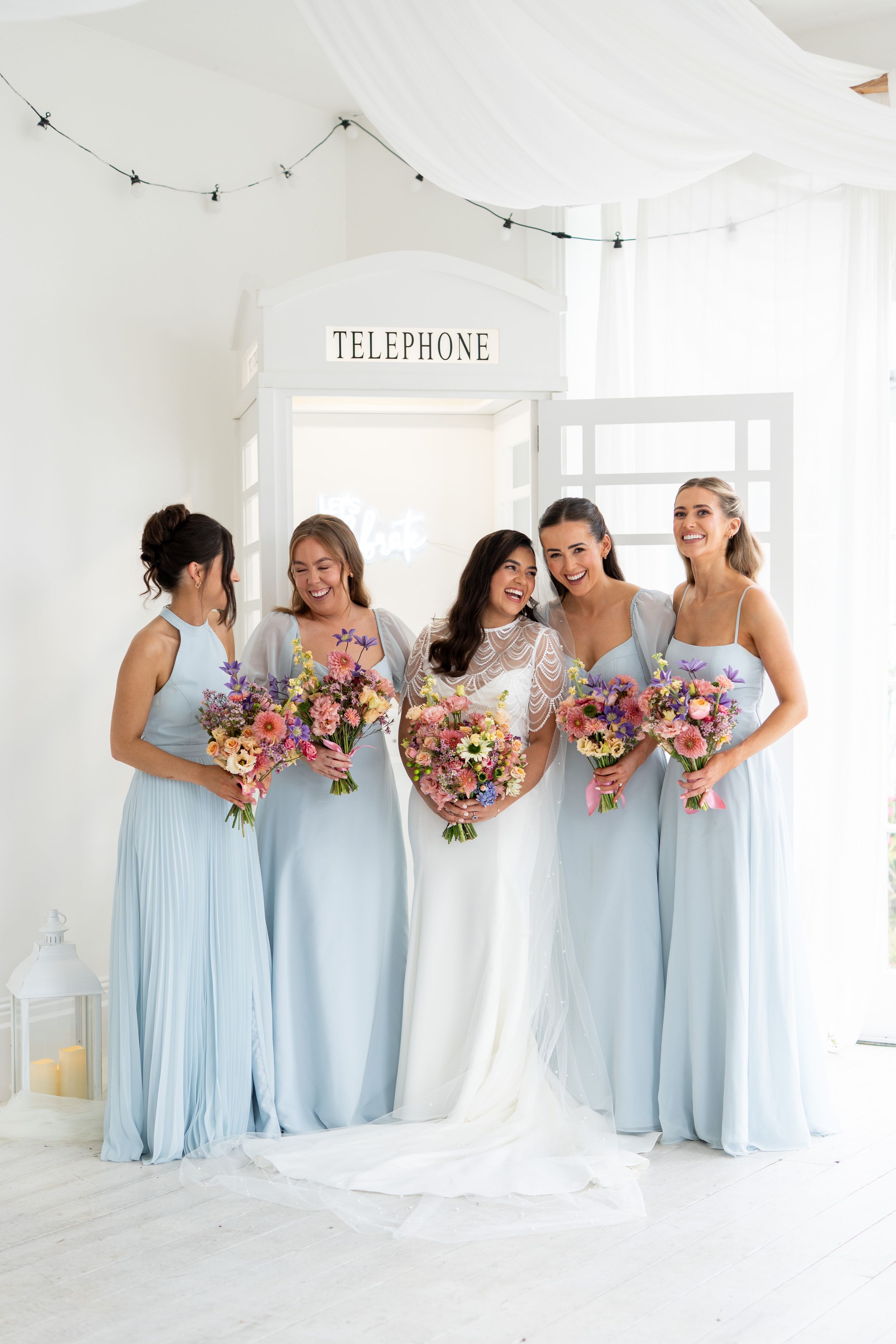 A group wearing matching light blue bridesmaid dresses and holding colorful bouquets pose together in a bright white room.