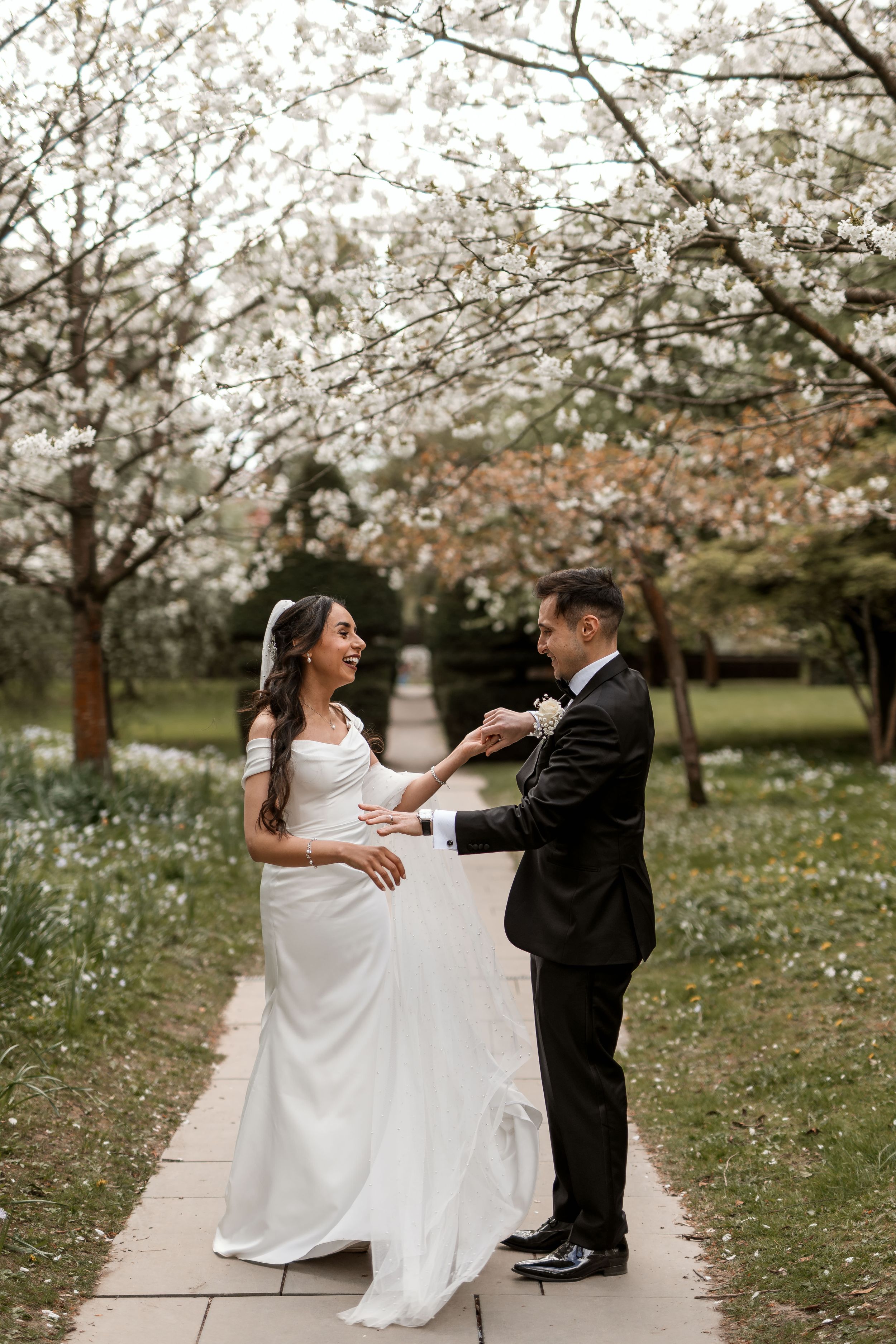 A couple in wedding attire dance and laugh together under blooming cherry blossom trees in a spring garden setting.