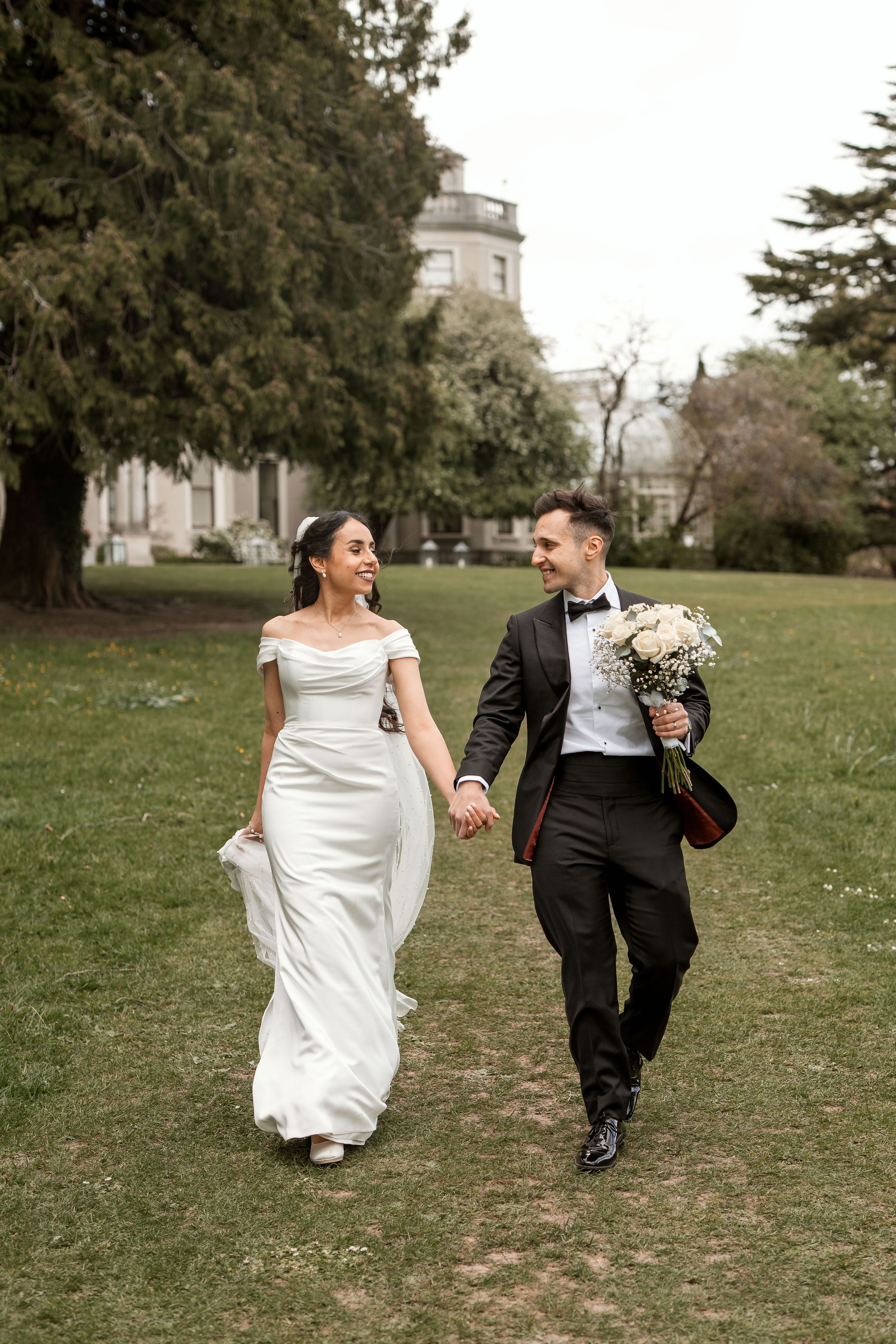 Happy newlyweds walking hand in hand on grass with white wedding dress and black tuxedo near historic building.