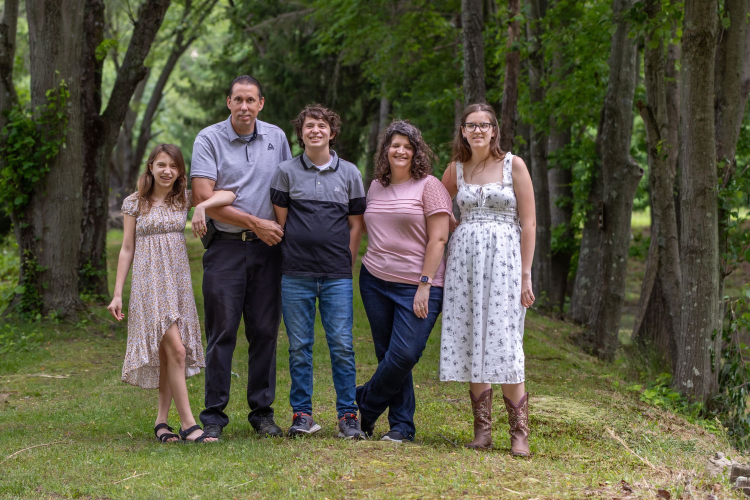 Family of five posing outdoors on green grass surrounded by tall trees in a casual summer photo session. Salem and Gloucester County Family