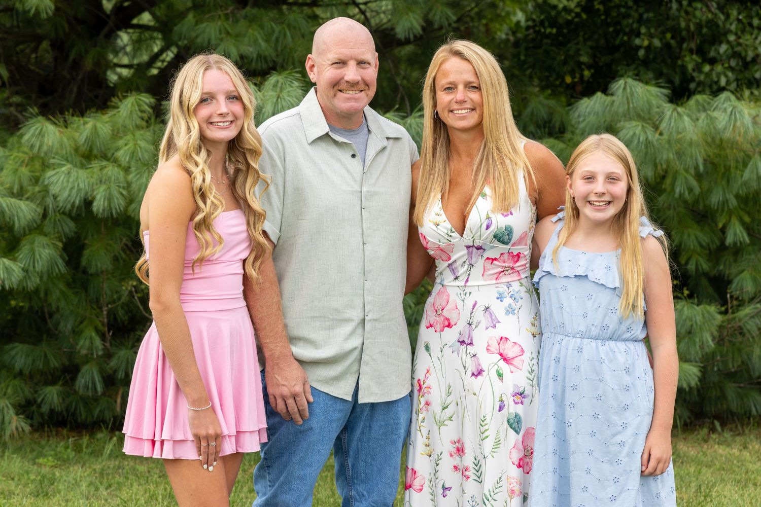 Family of four smiling outdoors, two blonde girls in pink and blue dresses with parents in a lush green setting. South Jersey Photographer