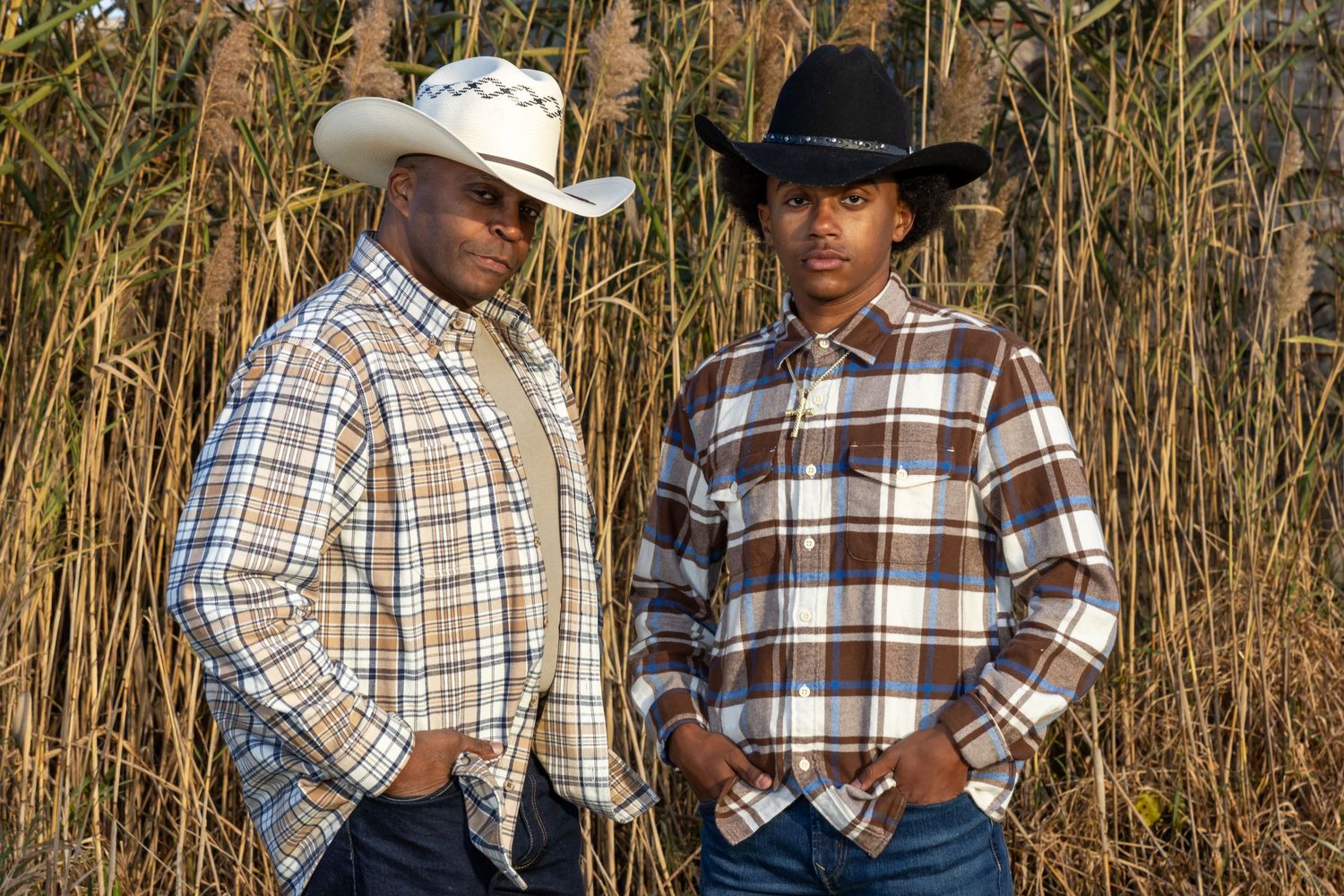 portrait of an African American father and son in plaid shirts and cowboy hats pose together against tall grass in a country setting.