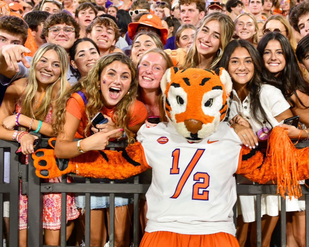 Enthusiastic students and fans pose with mascot wearing jersey at college sporting event.