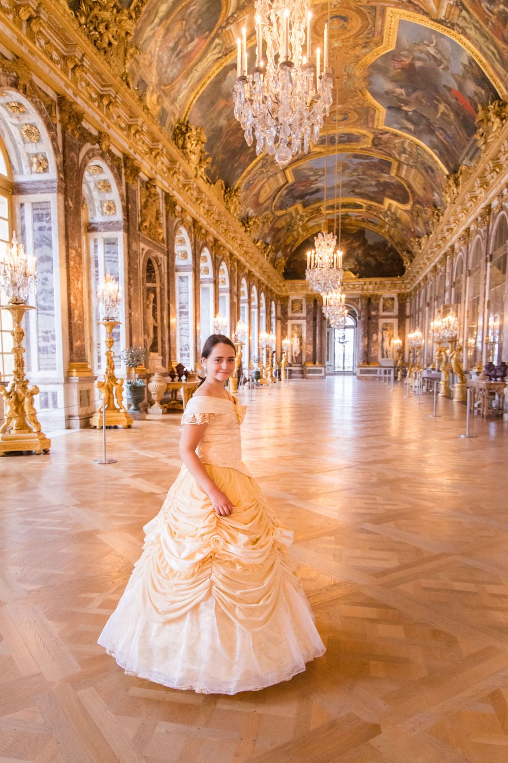 Woman in a gold gown posing in the Hall of Mirrors at the Palace of Versailles.