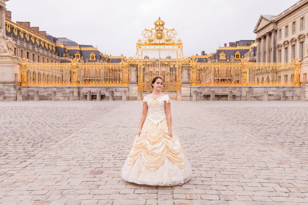 Woman in an elegant gown standing in front of the golden gates of the Palace of Versailles.