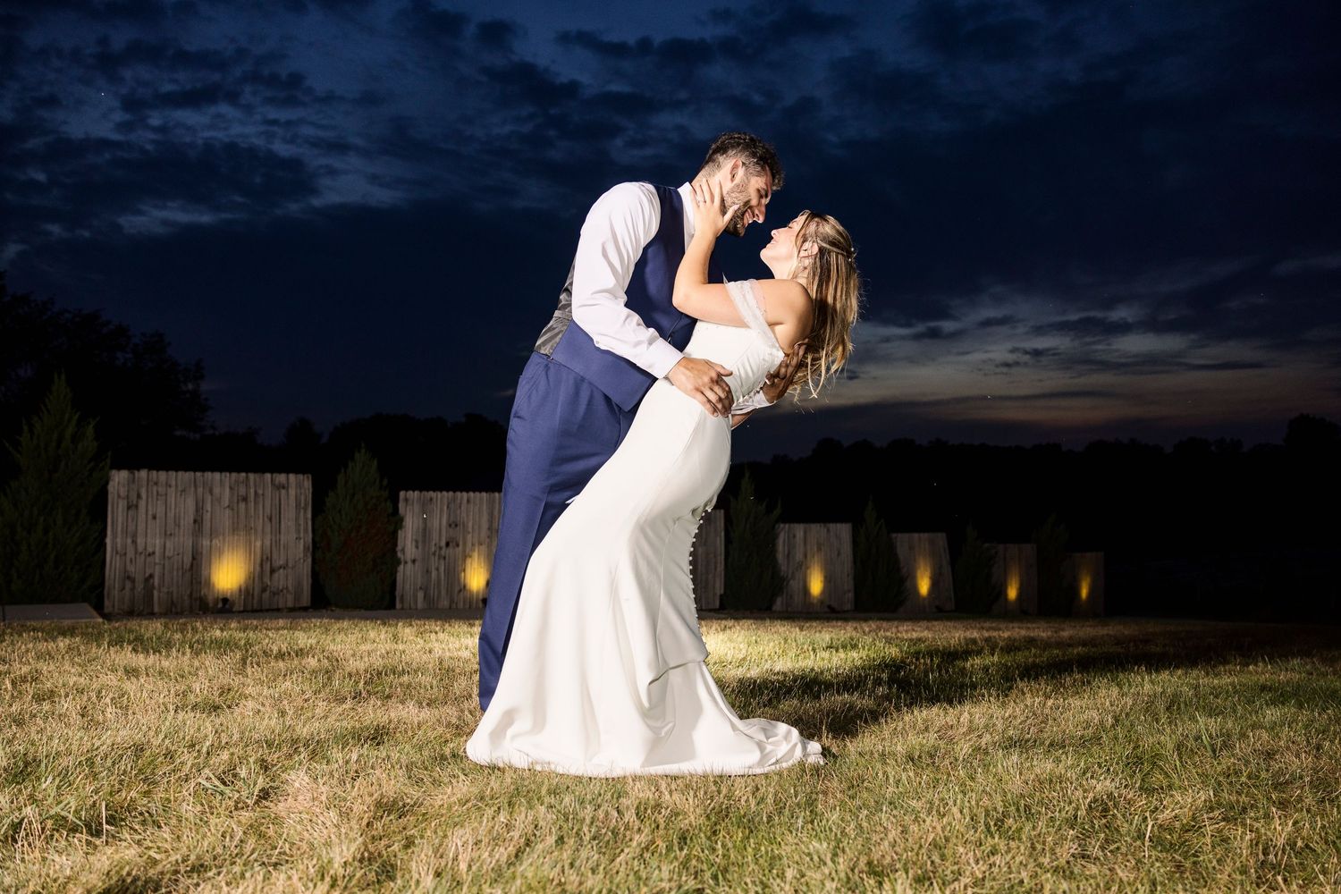 Bride and groom share a romantic dip kiss at night with glowing lanterns and dramatic blue sky backdrop.