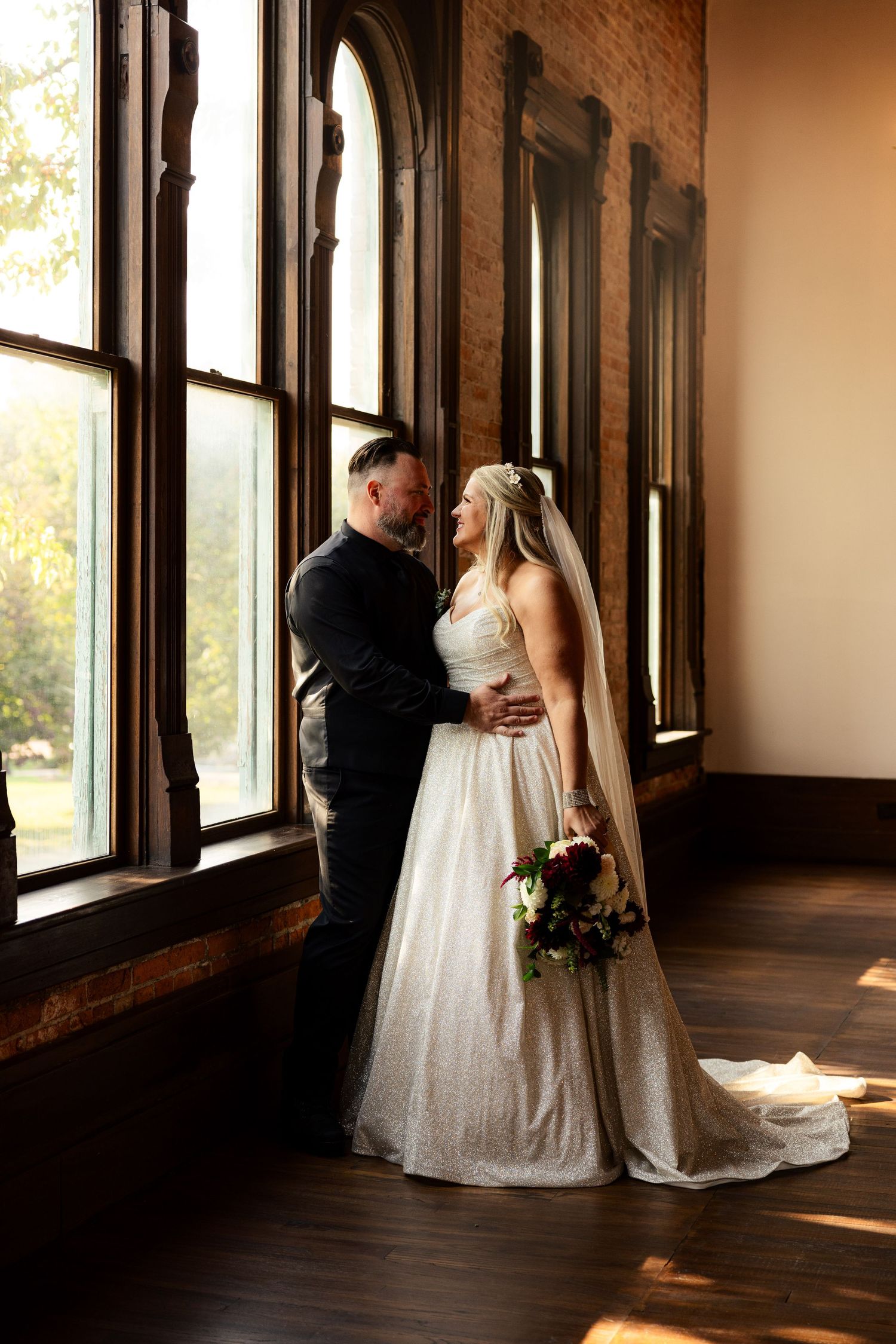 Bride and groom share a romantic moment by tall arched windows in a rustic brick wedding venue.