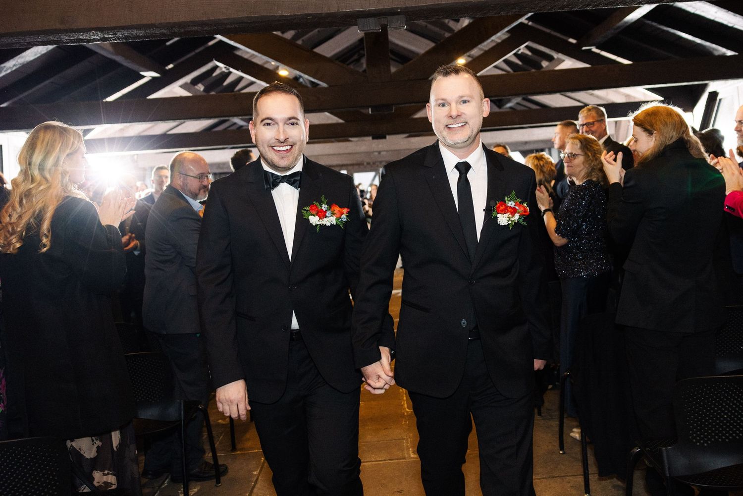 Two grooms in black tuxedos hold hands and smile while walking down the aisle at their wedding reception.