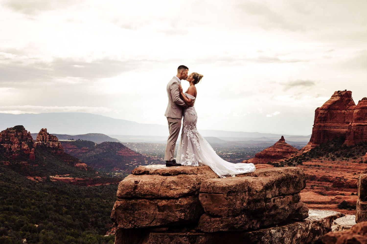 Bride and groom share a romantic kiss on a rocky cliff overlooking Sedona's stunning red rock landscape.