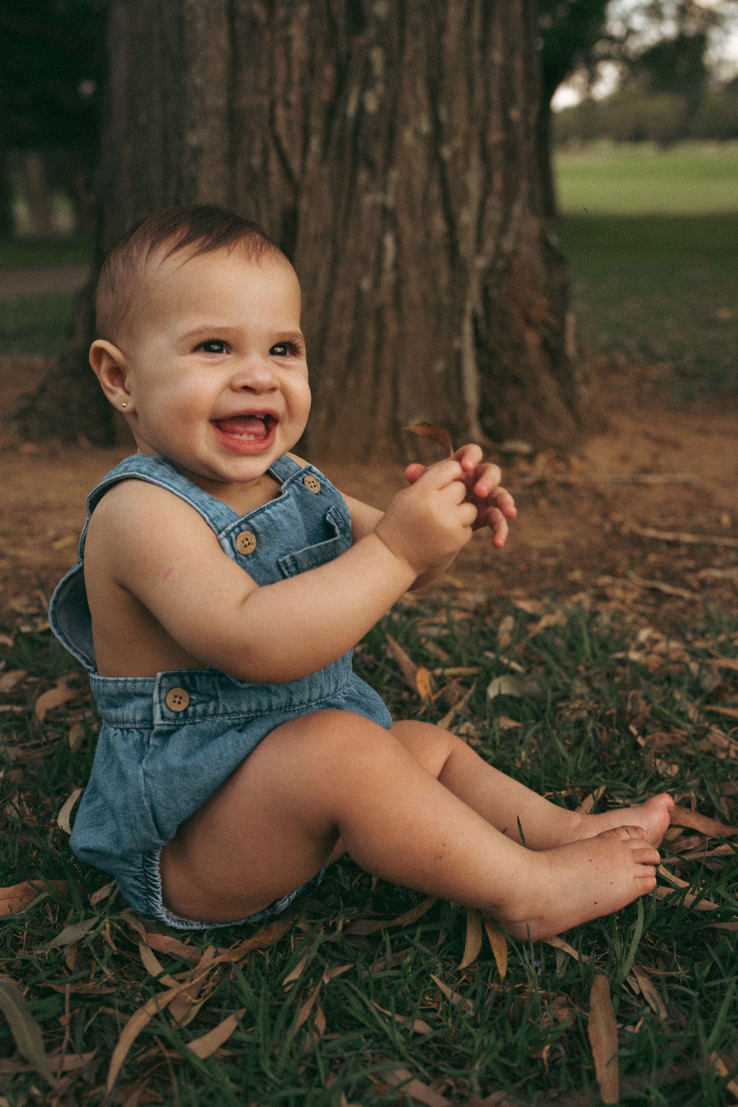 Baby in denim overalls sits playfully among fallen leaves near a tree trunk.